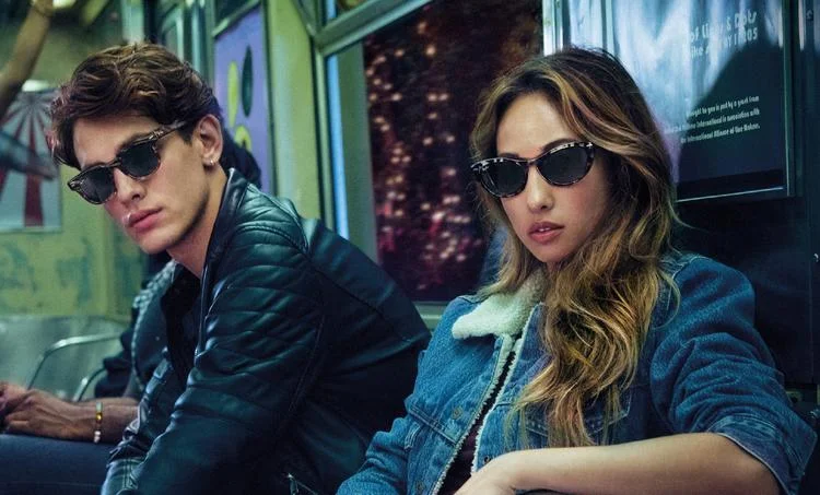 Two people in sunglasses and stylish jackets sit in a subway car, looking cool and relaxed, their Ray-Ban RB0707S Sunglasses from Ray-Ban with acetate frames adding an extra layer of chic.