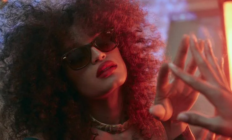 Person with curly hair and Ray-Ban MEGA WAYFARER RB0840S sunglasses making a hand gesture under red lighting.