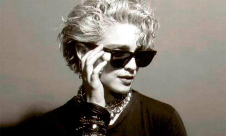 Black and white photo of a person with short, curly hair wearing Ray-Ban WAYFARER RB2140F eyeglasses, a dark shirt, and multiple necklaces.