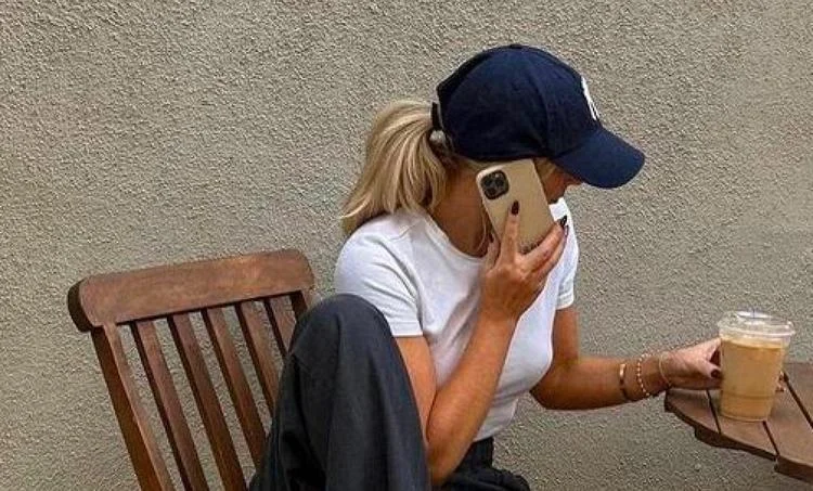 A person wearing a cap and Ray-Ban RB2210 Sunglasses, sitting on a chair, holding a phone to their ear and a cup of iced coffee.