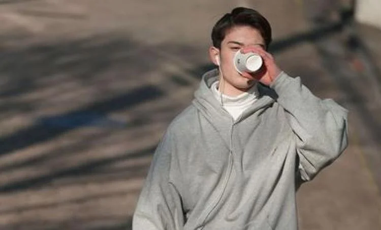 Person in a gray hoodie sipping from a paper cup while walking outside, wearing Ray-Ban RB2210 Sunglasses with polarized lenses, blurry shadows in the background.