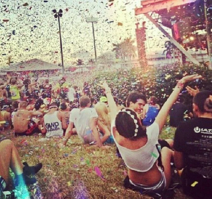 People sitting on the grass at an outdoor music festival, with confetti in the air and colorful lights shining. Many are wearing Ray-Ban BLAZE AVIATOR RB3584N sunglasses, enjoying the vibrant scenes with polarized lenses that make every detail pop.