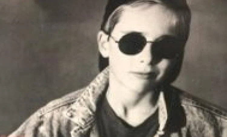 A black-and-white photo of a young boy wearing dark round Ray-Ban ELON RB3958 sunglasses, a cap backwards, and a denim jacket.