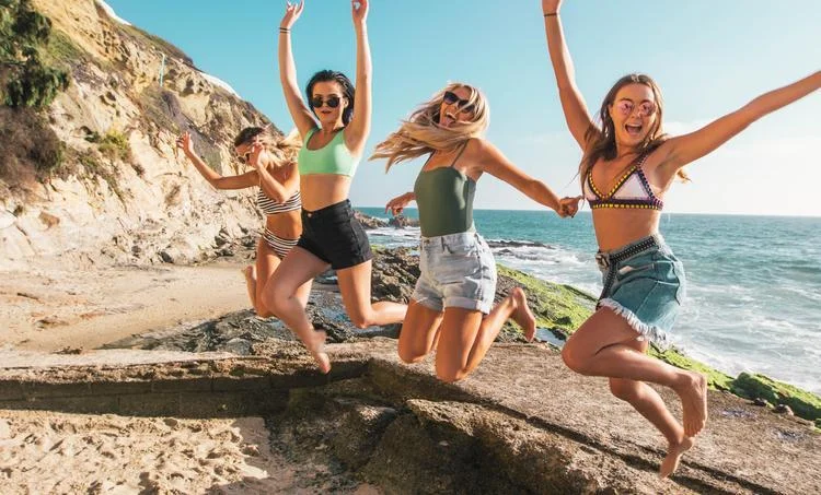 Four women in swimwear and Ray-Ban RB4337 Sunglasses joyfully jumping on a rocky beach by the sea with cliffs in the background.