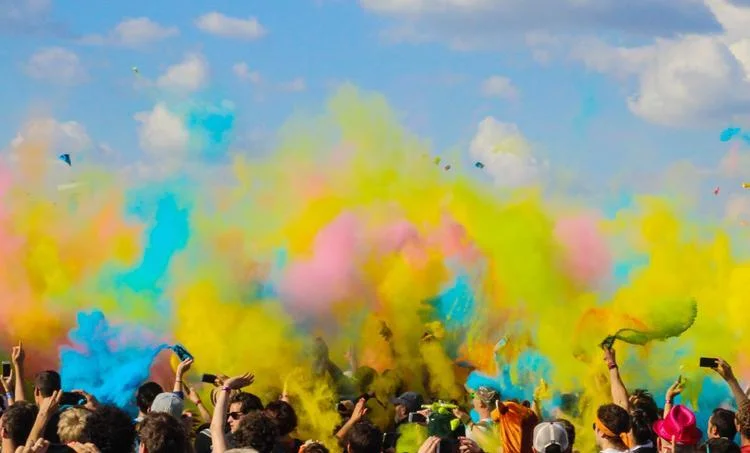 People in Ray-Ban RB4337 Sunglasses celebrate with vibrant colored powder in the air at an outdoor festival under a blue sky with scattered clouds.