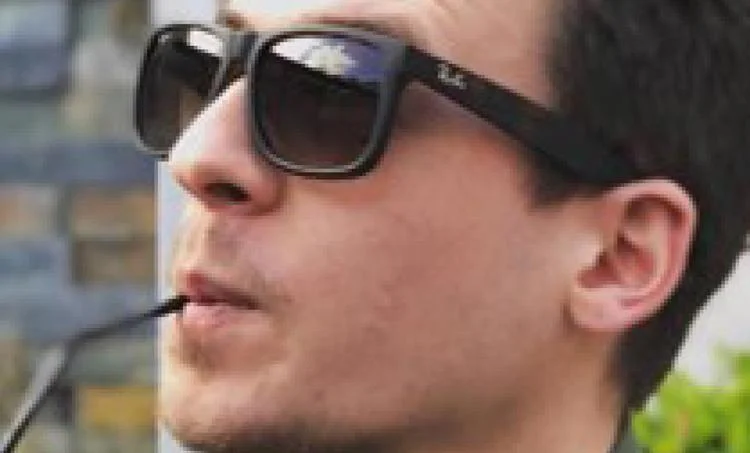 A person wearing Ray-Ban RB4362F Sunglasses | Size 55 with the tip of a minimal squared shape black frame in their mouth, looking up slightly. The background is blurred.