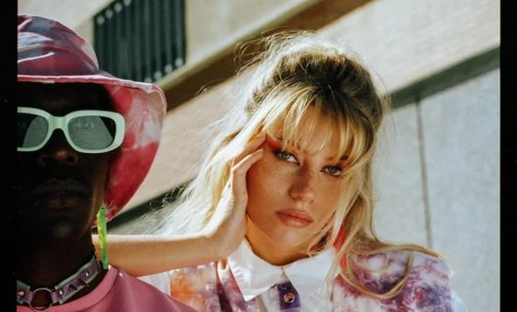 A person in Ray-Ban TERU RB4425 sunglasses and a bucket hat stands beside another individual posing with a hand to their temple, dressed in colorful tie-dye clothing, embracing a retro vibe.