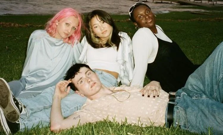 Four people pose together on the grass, some sitting and some lying down, all smiling at the camera in their Ray-Ban TERU RB4425 Sunglasses, radiating a retro vibe.