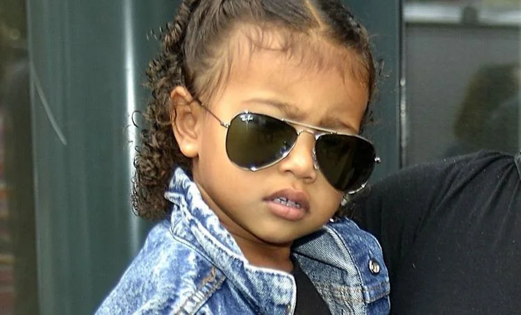 A young girl with curly hair wearing Ray-Ban JUNIOR STATE STREET RJ9186S sunglasses and a denim jacket, looking to the side with a serious expression.